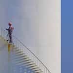 Engineer is walking up spiral staircase to working on top of fuel tank with blue sky background